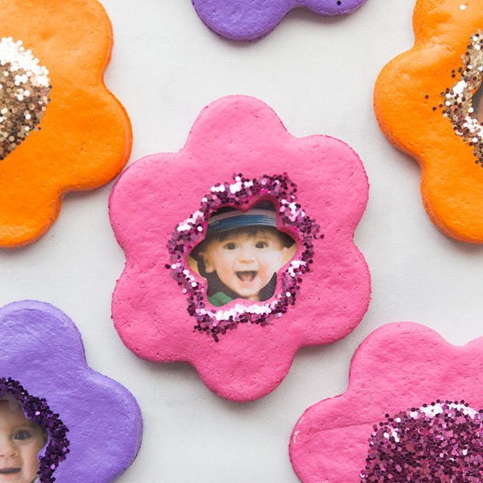 Colorful, flower-shaped decorations feature cut-out windows showcasing smiling children's photos, adorned with sparkling glitter accents, resting on a light surface.
