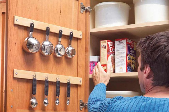 A person reaching for a box in a kitchen cabinet, while measuring cups hang on the inside of the cabinet door.