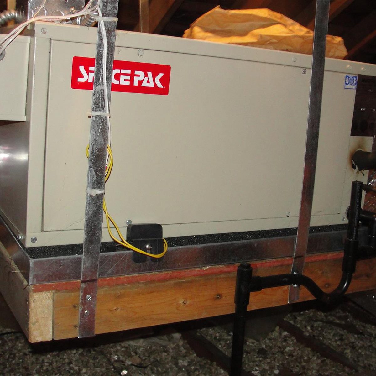 A large tan unit labeled "SPACEPAK" is positioned on a wooden platform, with visible electrical wires and pipes, in a dimly lit attic environment.