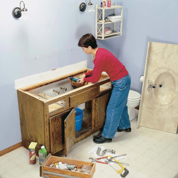 A person installs a sink in a bathroom, surrounded by tools and materials, with an open cabinet and a toilet in the background.