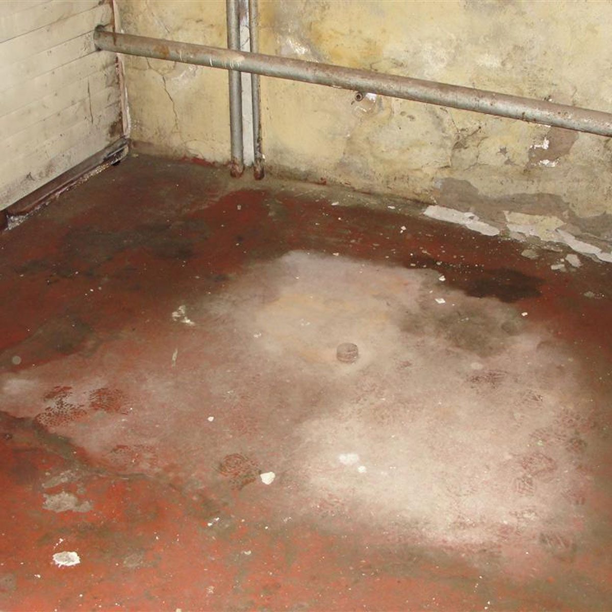 A stained concrete floor with patches of discoloration and footprints, surrounded by bare walls and exposed pipes in a dimly lit space.
