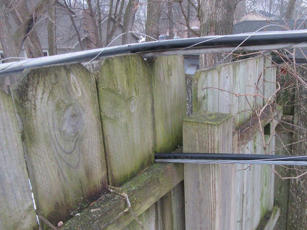 A wooden fence supports a black wire that stretches across its top, surrounded by bare trees and residential buildings in a cloudy environment.