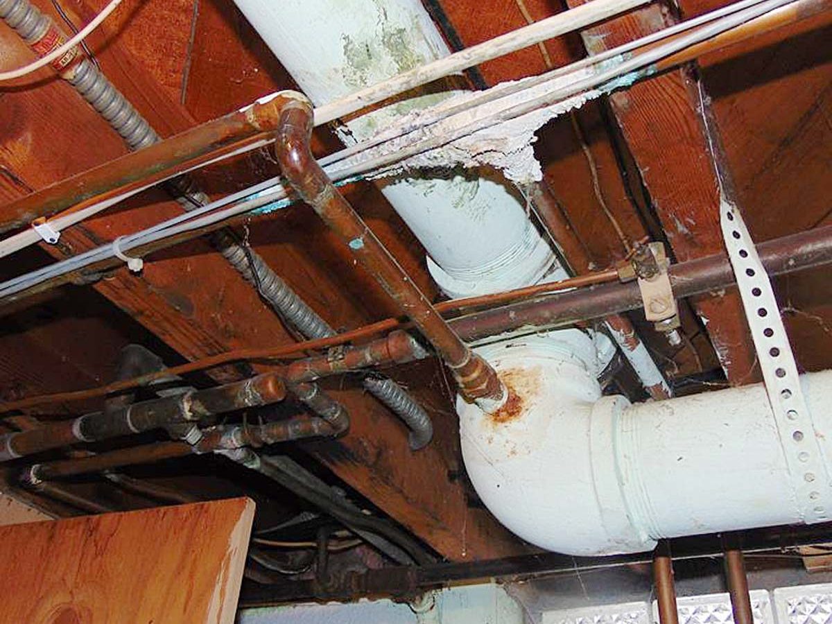 Pipes and ductwork traverse a wooden ceiling, with a white pipe turning at a joint, showing signs of wear. Light filters through a window.
