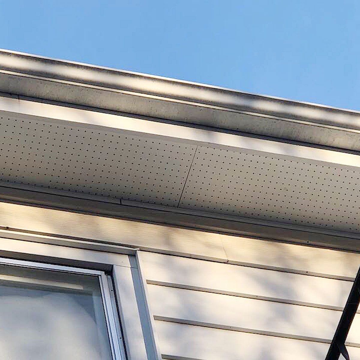 A house's eaves are visible, with perforated soffit panels installed beneath the roof. The sky is clear and blue in the background.