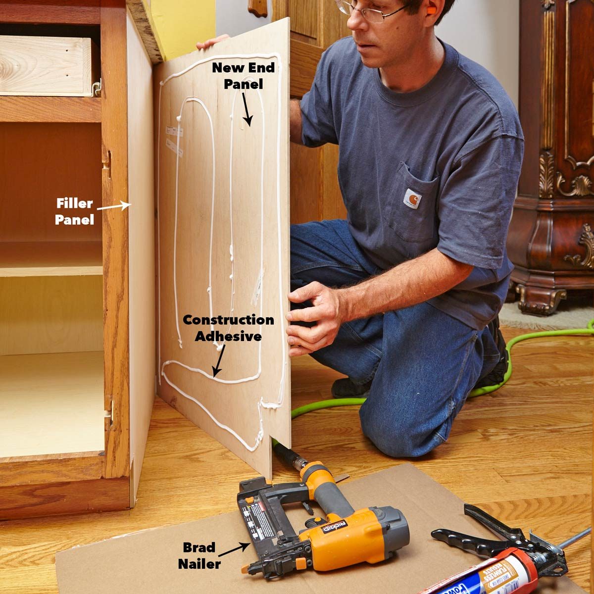 A man kneels, applying construction adhesive to a new end panel, preparing to install it alongside a filler panel on a kitchen cabinet.