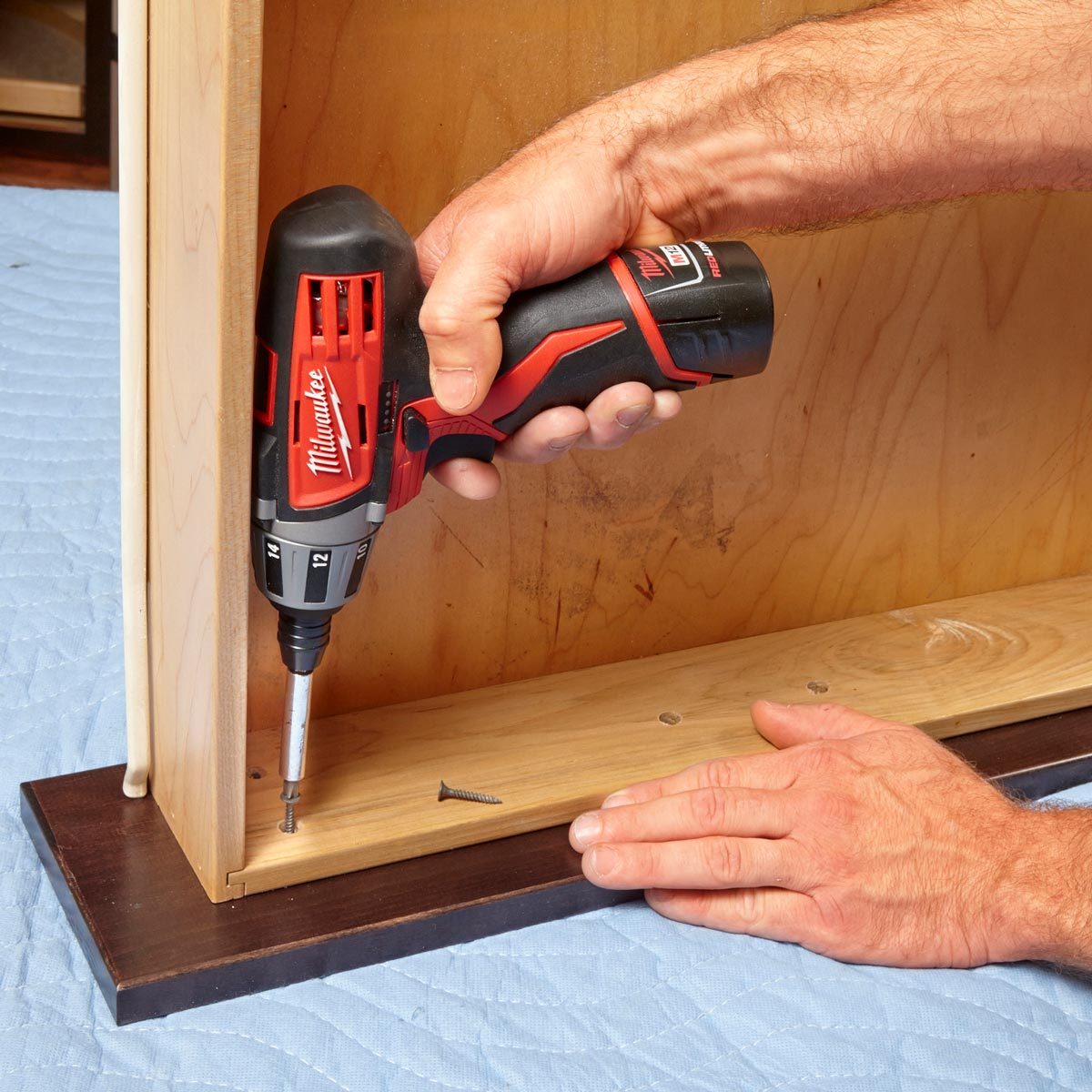 A person is using a power drill to screw into a wooden panel on a blue protective surface. Another hand stabilizes the panel.