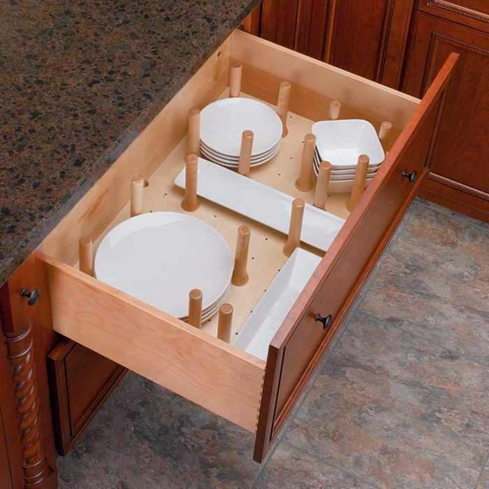 A kitchen drawer holds neatly arranged white plates and dishes, supported by wooden dividers, against a backdrop of dark countertop and tiled floor.