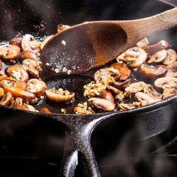 Sliced mushrooms are being sautéed in a cast iron skillet, stirred with a wooden spoon, surrounded by steam and cooking oil.