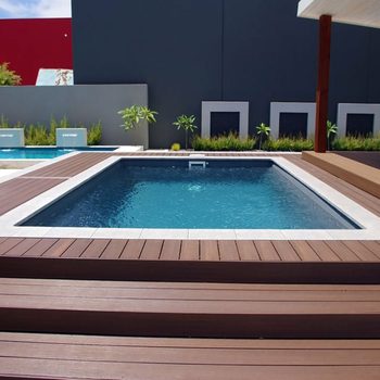 A rectangular swimming pool filled with clear water sits on wooden decking, surrounded by greenery and a modern, minimalist backdrop.