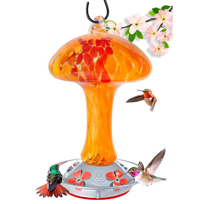 A vibrant, orange hummingbird feeder hangs from a hook, attracting multiple colorful hummingbirds while surrounded by delicate pink flowers.