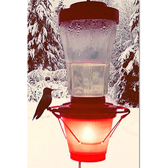 A red hummingbird feeder sits amidst snowy trees, with a bird perched on its edge, ready to drink from the top reservoir.