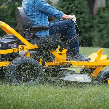 A riding lawn mower moves across a grassy area, cutting grass while ejecting clippings, with a person seated on it, focusing on lawn maintenance.