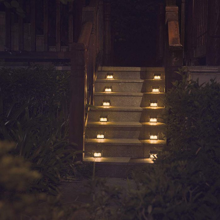 A staircase lights up with small glowing fixtures, illuminating each step at night, surrounded by greenery and wooden railings.