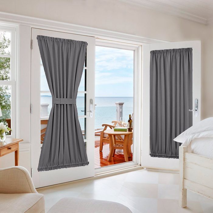 Two gray curtains hang on double doors leading to a seaside patio, where wooden chairs are visible against a backdrop of calm water and blue sky.
