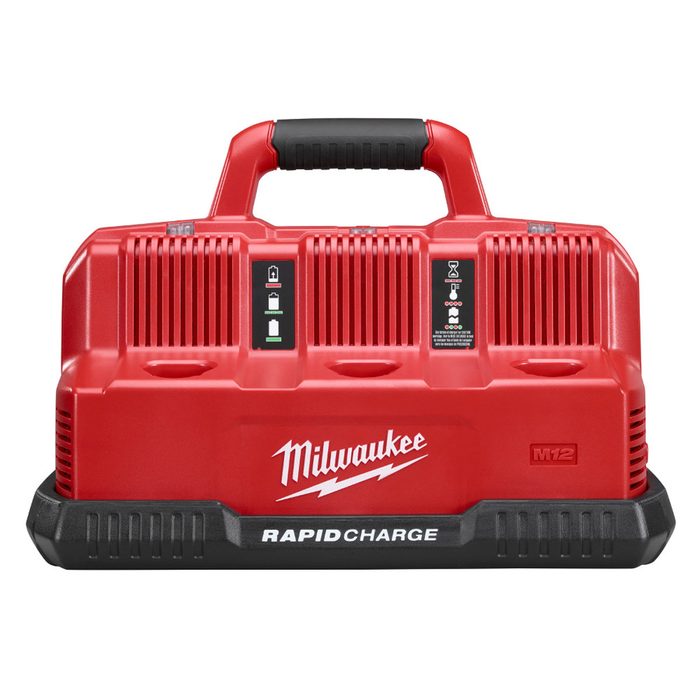 A red battery charger with a handle is positioned upright, displaying charging indicators and branding, designed for Milwaukee M12 batteries in a tool-oriented context.
