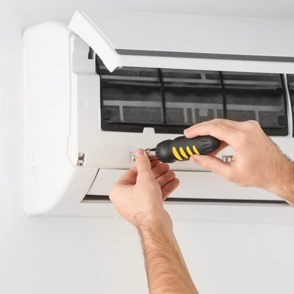 A person uses a screwdriver to remove a cover from an air conditioning unit, surrounded by a white wall and the unit itself.
