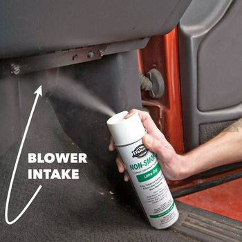 A person sprays cleaning solution into a vehicle's blower intake, which is located beneath the dashboard. The environment appears to be the interior of a car.