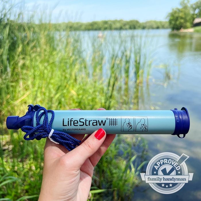 A person holds a LifeStraw water filtration device by a lake, surrounded by tall grasses and trees under a clear blue sky.