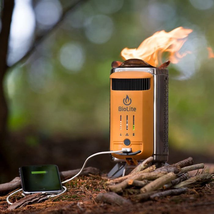 A portable stove emits flames while charging a phone, surrounded by sticks on the forest floor under soft, diffused light.