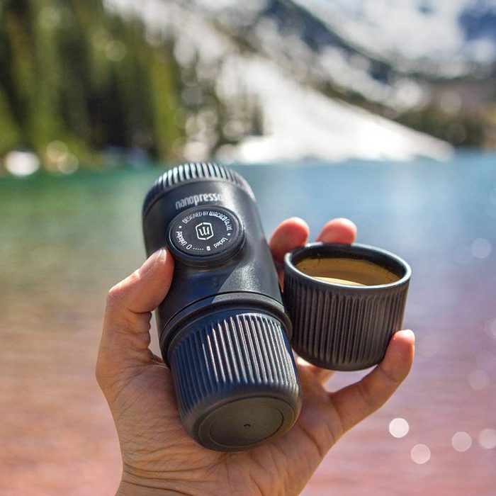 A black coffee maker is held in one hand, releasing a small cup of coffee, with a scenic lake and mountains in the background, indicating outdoor use.