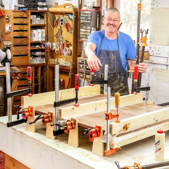 A man holds clamps while adjusting wood pieces on a workbench, surrounded by tools and organized storage in a workshop environment.
