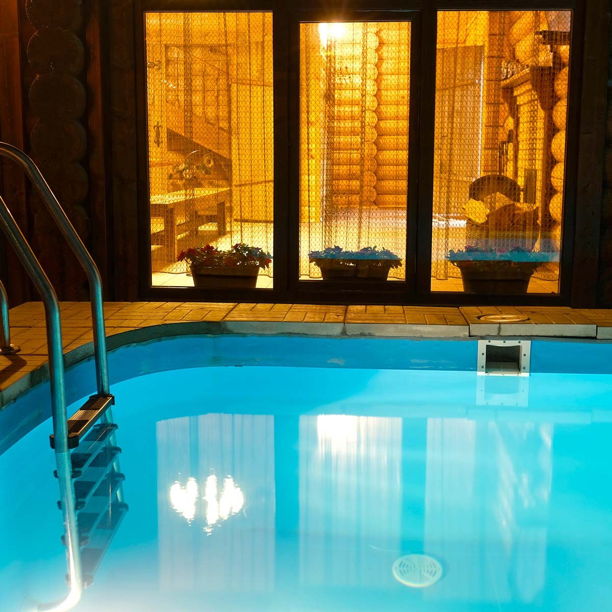 A swimming pool with a ladder reflects overhead lights, situated beside large glass windows revealing a warm, cozy interior with potted plants.