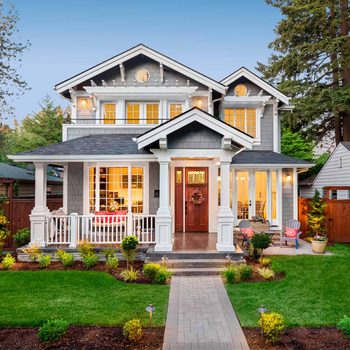 A modern house with a grey exterior features a welcoming porch, large windows, and a pathway leading to the front door, surrounded by green lawn and flowers.