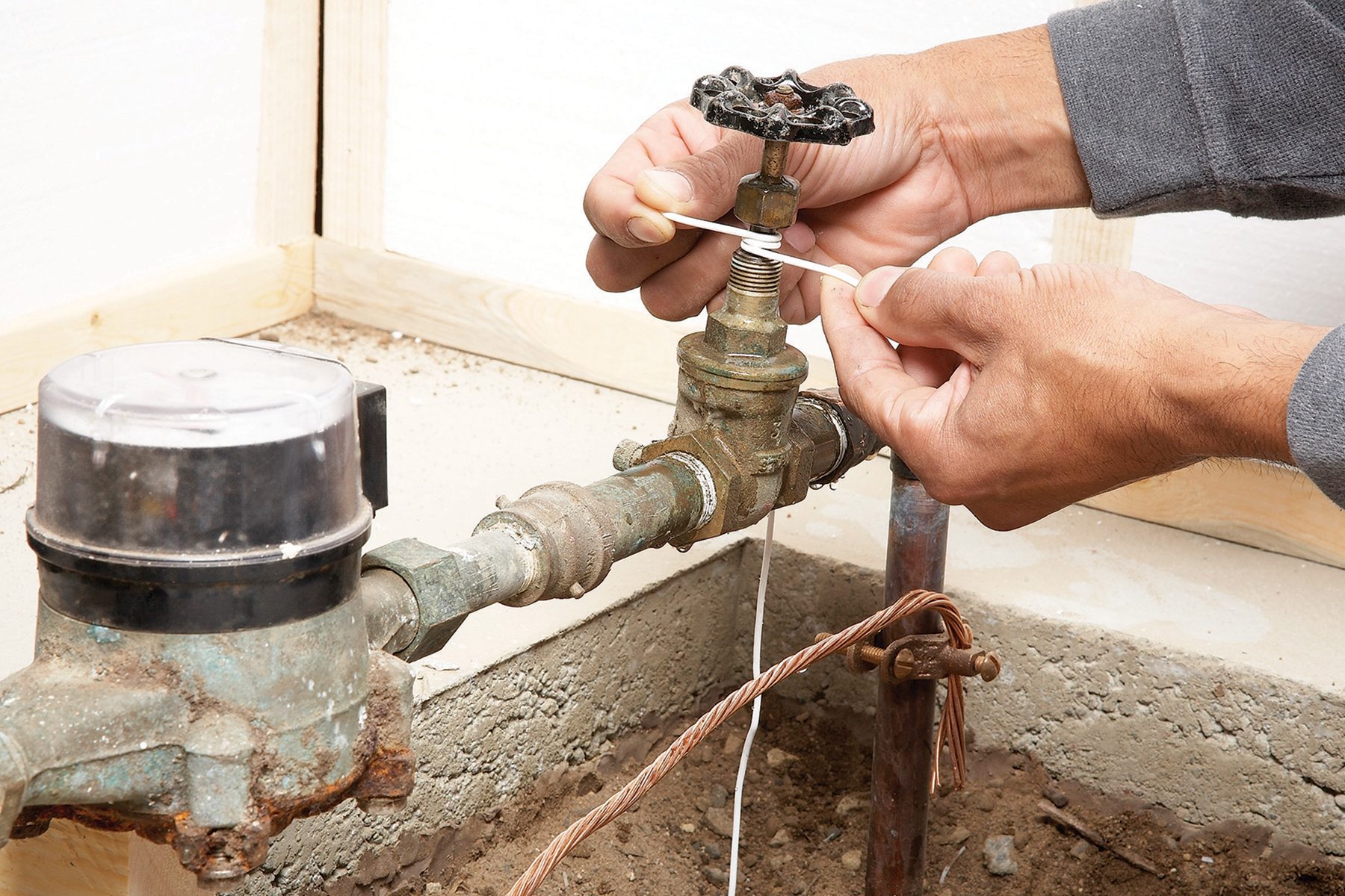 A person tightens a valve using a rubber band on a water meter and pipe system, situated in a dirt-floored area with wooden framing.