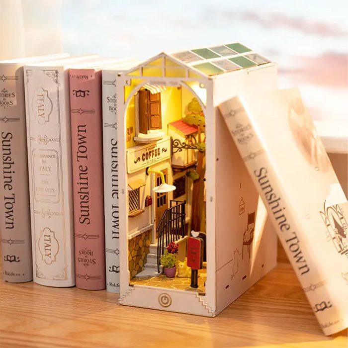 A miniature coffee shop nestled between books, illuminated from within, with stairs, greenery, and a small detail of a cat outside its welcoming entrance.