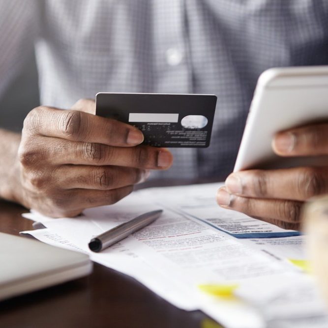 Close-up view of African man's hands holding plastic credit card and smart phone. Dark-skinned businessman using mobile banking application on his gadget while paying bills at cafe online via internet