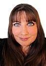 A woman with long brown hair and bangs is smiling at the camera against a plain white background. Her eyes appear bright and expressive.