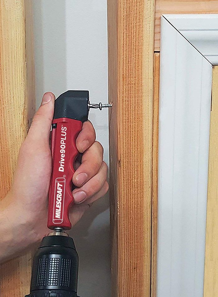A hand holds a red tool driving a screw into wooden framing in a bright indoor setting.