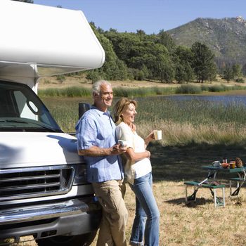 A couple stands by a camper van, holding cups. They smile while leaning against the vehicle, with a serene lake and mountains in the background.