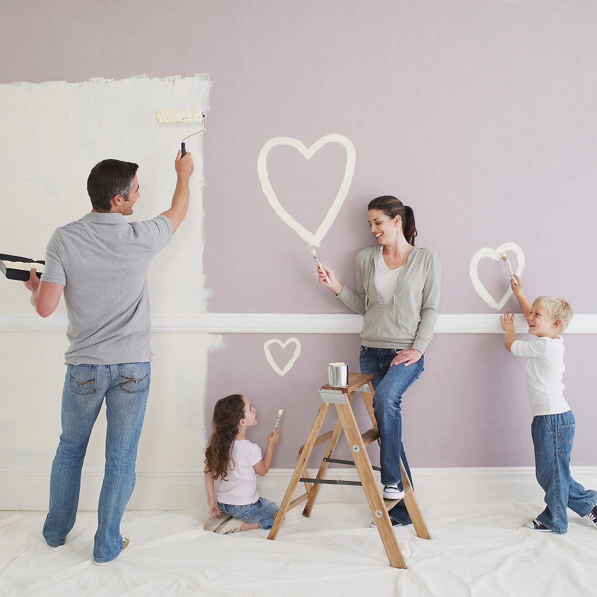 A family paints a wall, applying light colors and heart shapes. The scene is indoors, with a ladder and drop cloth on the floor.