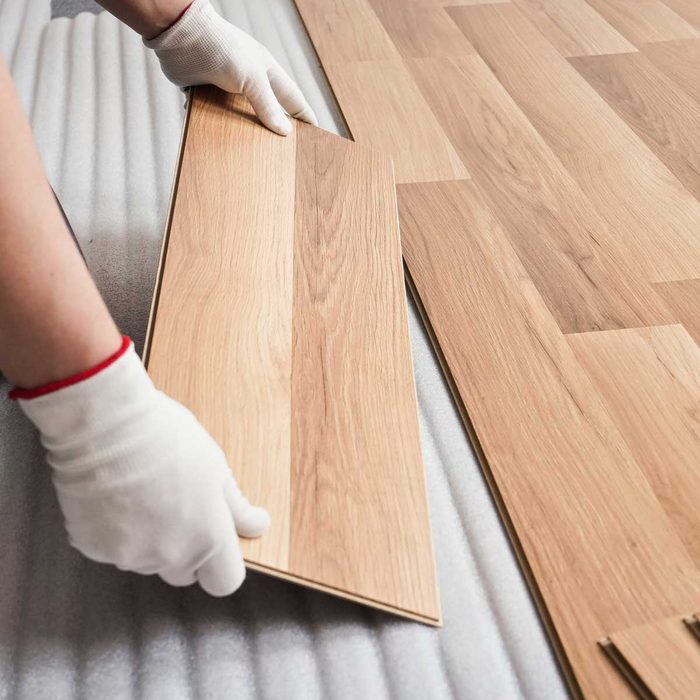 A gloved hand is placing a wooden flooring panel onto a padded surface, surrounded by additional panels arranged in a room undergoing installation.