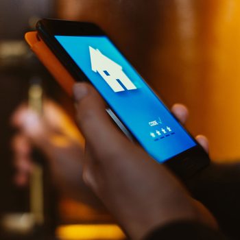 A hand holds a smartphone displaying a blue screen with a house icon. The background is blurred, hinting at an indoor setting.