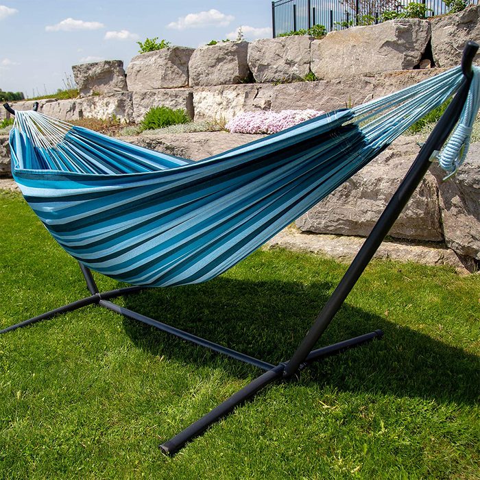 A striped blue hammock hangs from a black stand, resting on green grass, surrounded by stone landscaping and under a clear blue sky.
