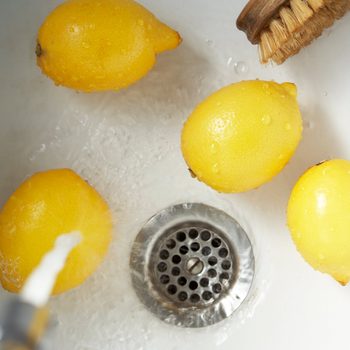 Yellow lemons are submerged in running water within a sink, surrounded by droplets and a metal drain. A brush is visible nearby.