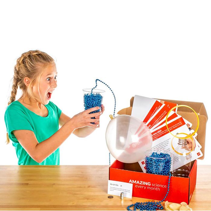 A girl, excited and surprised, holds a cup filled with blue beads; a science kit box and a balloon are nearby on a wooden table.