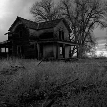 A dilapidated wooden house stands in overgrown grass, weathered by time, under a cloudy sky, surrounded by bare trees.