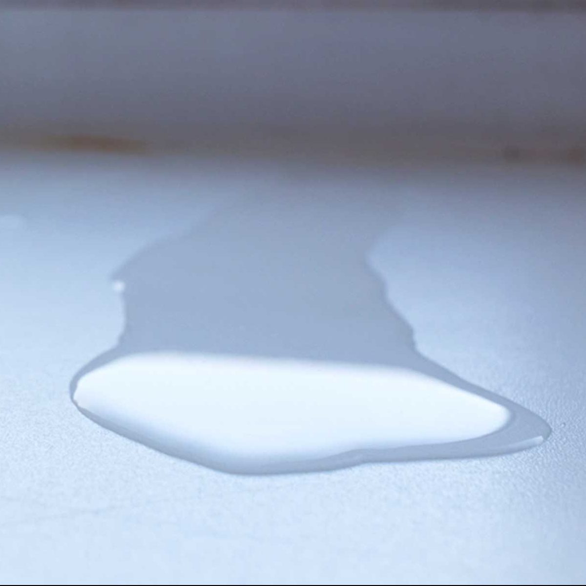 A puddle of clear liquid rests on a flat, light-colored surface, reflecting light, suggesting recent spillage in a clean indoor environment.