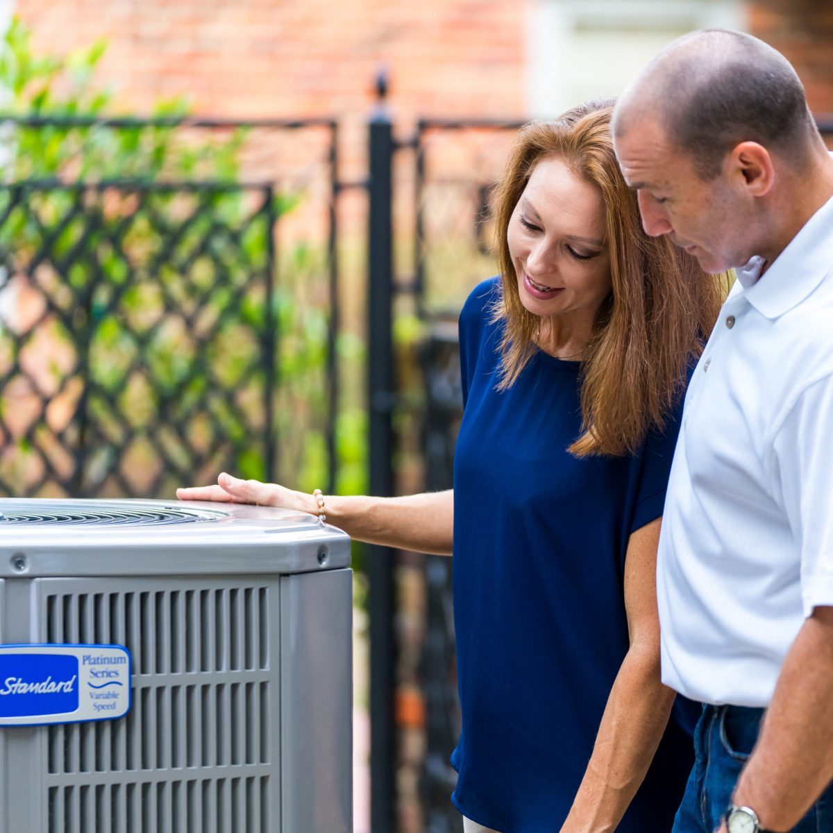 A woman and a man inspect an outdoor air conditioning unit, engaged in conversation, with a green, textured background of foliage and a fence.