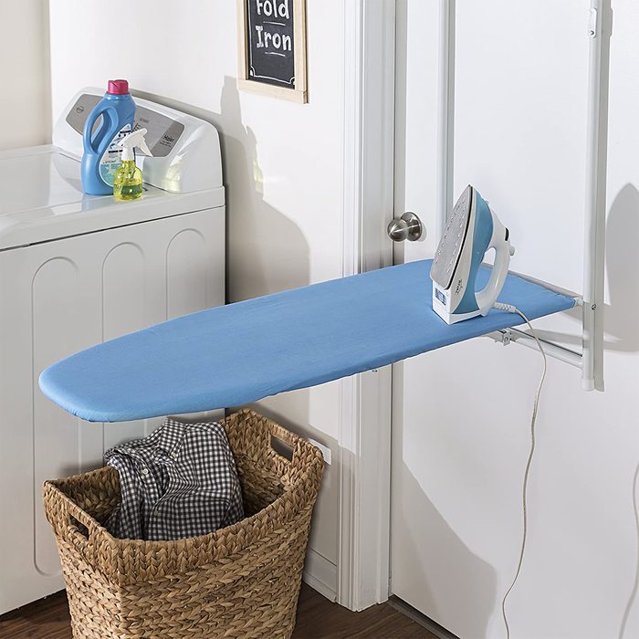 An ironing board with a blue cover holds an iron, positioned near a washing machine and a laundry basket filled with clothes.