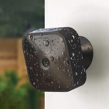 A black security camera, covered in water droplets, is mounted on a white surface, observing a blurred, lush green background.