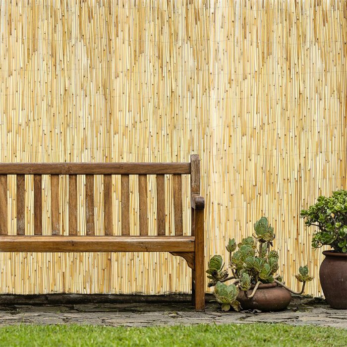 A wooden bench sits beside potted plants against a textured bamboo wall in a grassy outdoor setting, creating a serene and inviting atmosphere.