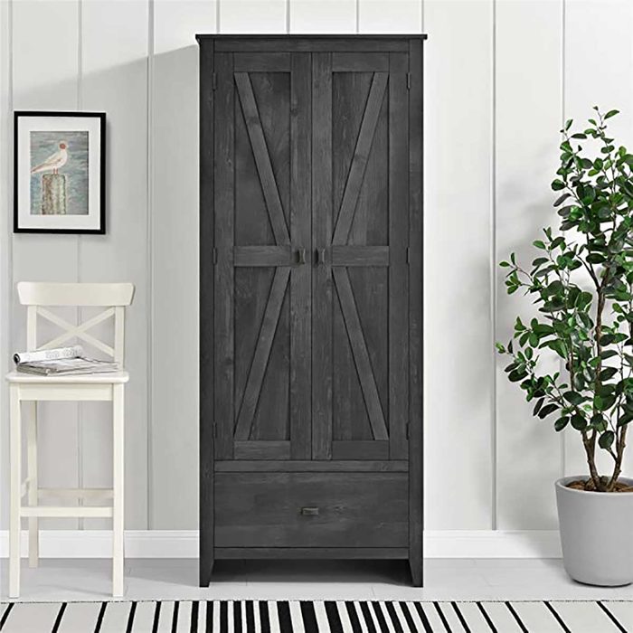 A tall, dark wooden wardrobe stands against a light wall, accompanied by a white chair and a potted plant, creating a cozy interior setting.