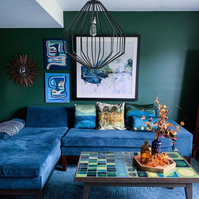 A blue couch with decorative pillows sits beneath a decorative light fixture, surrounded by vibrant wall art in a warmly colored room.