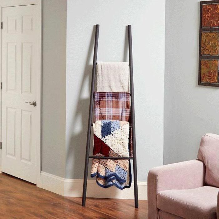 A ladder-shaped rack holds folded blankets in a corner of a room, beside a light-colored couch and a door, enhancing the cozy ambiance.