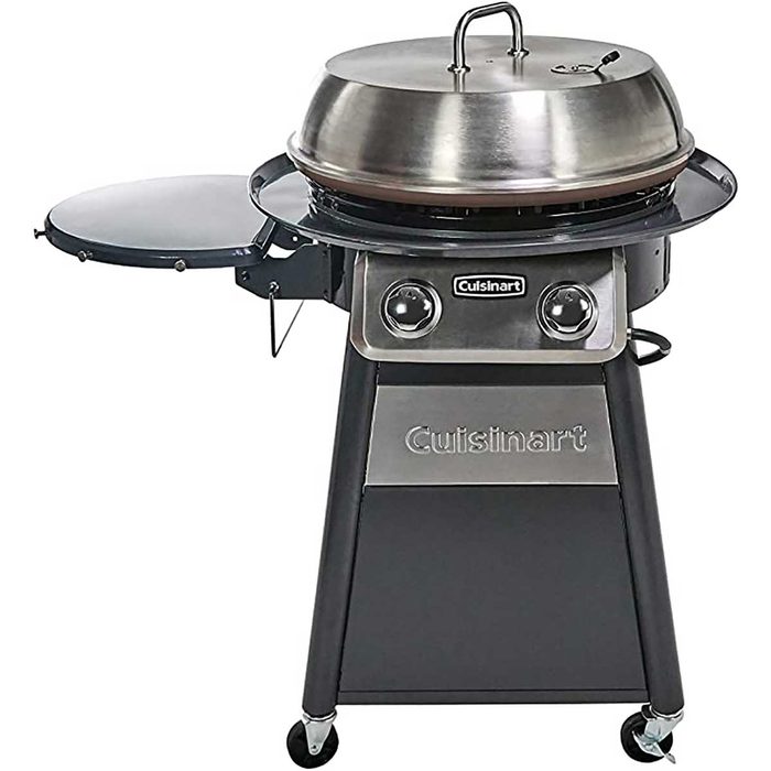 A round, stainless steel grill with a dome lid is set on a sturdy cart. It features two burners and a side table for food preparation.