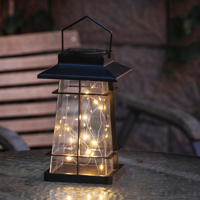 A decorative lantern filled with glowing string lights sits on a glass table in a dimly lit outdoor setting, with wooden benches visible in the background.